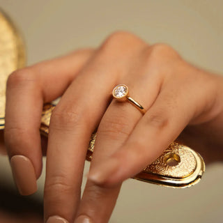 a woman's hand holding a bronze colored hand mirror wearing a yellow gold lab grown diamond engagement ring with a bezel setting on her ring finger
