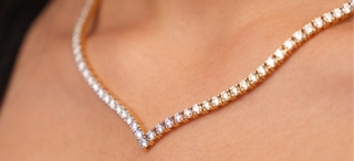 A close-up of an elegant, V-shaped gold necklace set with a single row of sparkling diamonds, resting on a woman's collarbone