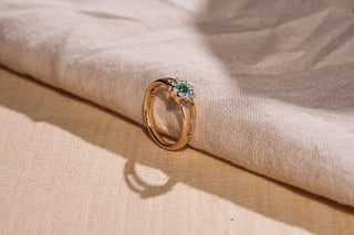 A gold ring with a floral cluster of a central emerald and surrounding lab-grown diamonds rests on draped beige fabric.