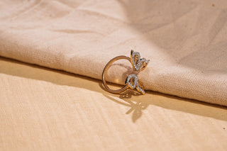 A gold open ring with two butterfly wings set with lab-grown diamonds rests on draped beige and tan fabric.