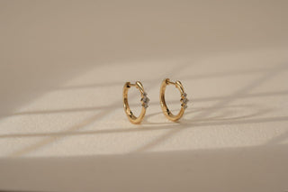 Pair of small yellow gold huggie hoop earrings, each set with three flush-set lab-grown diamonds, resting on a light beige surface.