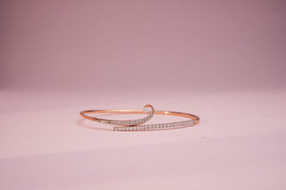 Rose gold cuff bangle with a swirling design, featuring channel-set lab-grown diamonds and a solitaire accent, on a light pink background.