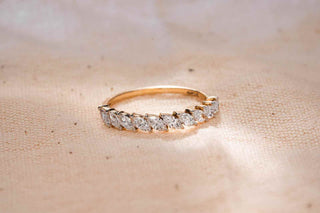 A gold band ring featuring a row of sparkling, round-cut diamonds set in a continuous line, placed on a soft beige fabric with gentle light reflections highlighting the brilliance of the stones