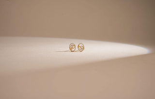 Pair of yellow gold stud earrings with a swirling design partially set with pavé lab-grown diamonds, resting on a light beige surface