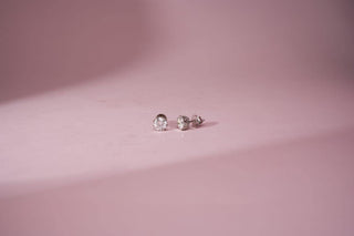 A pair of simple diamond stud earrings on a light pink background, with soft lighting and subtle shadows.