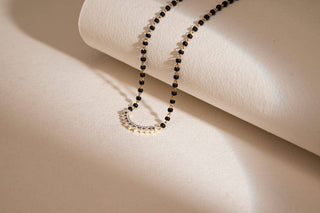 "A delicate mangalsutra necklace with small black beads and a semicircular pendant adorned with tiny white stones, displayed on a soft beige textured background with gentle lighting