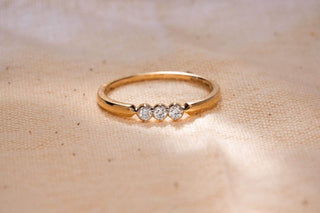 A close-up of a delicate gold ring with three small diamonds set across the top, resting on a textured fabric surface.