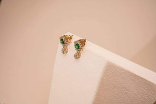 A pair of gold stud earrings with green gemstone centers and dangling white stones, elegantly displayed on a textured white geometric surface with a warm-toned background.
