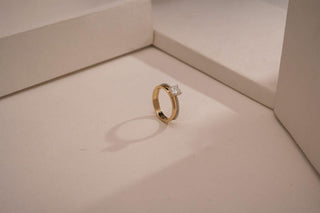 A gold engagement ring with a prominent square-cut diamond is standing upright on a light-colored surface, casting a soft shadow. The background consists of minimalist geometric shapes and clean lines, giving the scene a modern, elegant look.