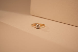 A simple gold ring featuring a single round-cut diamond in a classic solitaire setting, placed on a beige surface with soft, warm lighting creating gentle shadows.