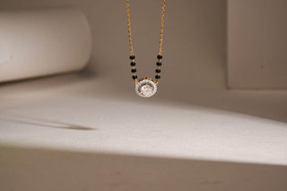 A delicate gold necklace featuring a central round diamond pendant, flanked by small black beads on each side, is suspended in front of a minimalist background with soft lighting and neutral-toned surfaces.