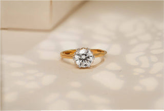 Gold solitaire ring with a large round diamond, displayed on a soft beige surface with shadow patterns