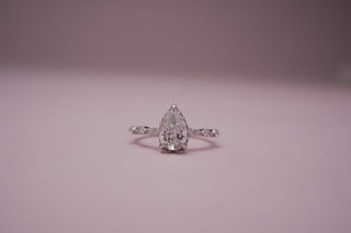 Silver ring with a pear-shaped diamond centerpiece, displayed on a soft pink background.