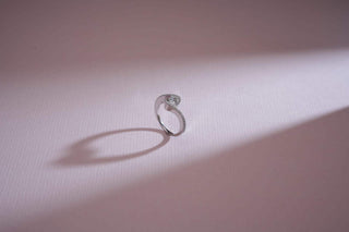 A white gold bypass ring with a swirling lab-grown diamond cluster stands on a soft pink surface."