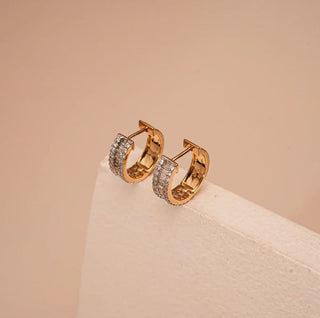 A pair of yellow gold huggie hoop earrings featuring baguette and round lab-grown diamonds rests on a white block
