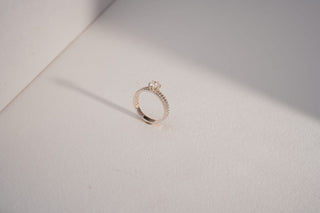A delicate rose gold  ring with a round central lab-grown diamond and double pavé bands stands on white.
