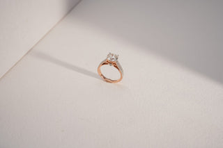 A rose gold ring with a large central lab-grown diamond and a pavé-set, split-shank band stands on a white surface.