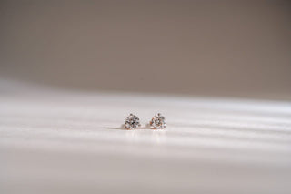 A pair of round brilliant lab-grown diamond stud earrings in white gold three-prong settings on a white surface.