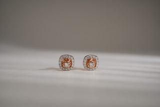 Rose and white gold lab-grown diamond stud earrings with a halo setting in a cushion-like shape are displayed on a light gray surface.