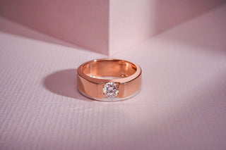 A rose gold band with white gold trim holds a round lab-grown diamond in a tension setting, on a pink textured surface.