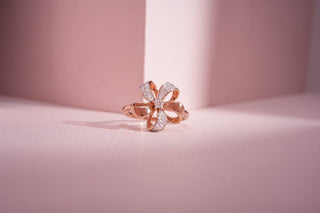 A rose gold lab-grown diamond ring is designed as a flower/bow with pavé-set stones and rests on a textured light pink surface.