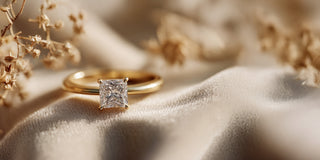 A Reia yellow gold engagement ring with a princess cut lab-grown diamond is displayed on folded beige velvet near small dried flowers.