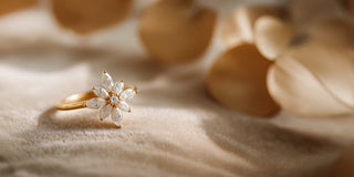 A yellow gold lab-grown diamond ring shaped like a flower rests on soft beige fabric near dry leaves.