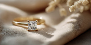 A Reia yellow gold engagement ring with a princess cut lab-grown diamond is displayed on folded beige velvet near small dried flowers.