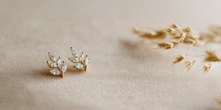 Yellow gold lab-grown diamond stud earrings, shaped like leafy branches, rest on beige fabric next to dried wheat stems.