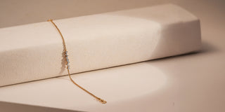 A thin gold chain bracelet with small lab-grown diamonds rests on a block, against a beige background.