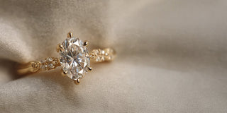 A Réia yellow gold engagement ring with an oval lab-grown diamond in a six-prong setting and diamond accents on the band, resting on beige fabric.