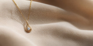 Reia yellow gold necklace with a subtle, twisting teardrop pendant. The pendant holds a single, round lab-grown diamond, resting on soft, flowing beige fabric.