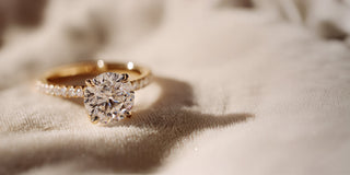Réia yellow gold solitaire ring with a pavé lab-grown diamond band, resting on beige fabric.