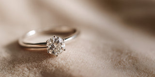 A Reia white gold solitaire engagement ring features a six-prong setting holding a round brilliant lab-grown diamond, resting on textured beige fabric.