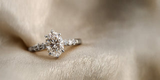 A Reia white gold lab grown diamond engagement ring features an oval lab-grown diamond in a six-prong setting, with small diamonds set on the band, resting on satin fabric.