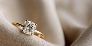 A Réia yellow gold solitaire engagement ring with a Cushion-cut lab-grown diamond in a four-prong setting, displayed on flowing satin fabric.