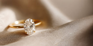 A Reia yellow gold solitaire engagement ring featuring a large oval lab-grown diamond in a four-prong setting, displayed on satin fabric.