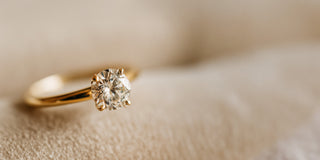 A yellow gold lab-grown diamond solitaire ring features a round brilliant cut stone in a six-prong setting, resting on soft beige fabric.