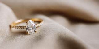 Réia yellow gold ring featuring a pear-cut lab-grown diamond on a pavé band, set on beige fabric.