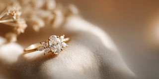 A Réia yellow gold ring with an oval lab-grown diamond center stone and marquise lab-grown diamond side accents, resting on beige fabric with dried flowers.
