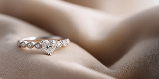 A white gold lab-grown diamond ring features a heart-shaped center stone with smaller accents set in bezels, on folded beige silk
