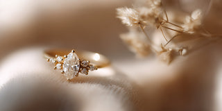 A Réia yellow gold engagement ring with a marquise-cut lab-grown diamond center stone and diamond accents, resting on beige fabric with dried flowers.