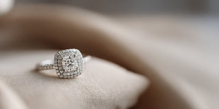 A Réia white gold engagement ring featuring a square center diamond with a double diamond halo and pavé band, resting on beige fabric.