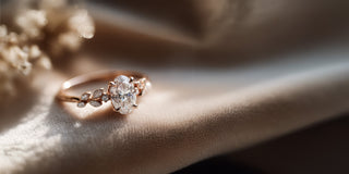 A Réia rose gold ring with an oval lab-grown diamond center stone and marquise and round diamond floral accents, resting on beige fabric.