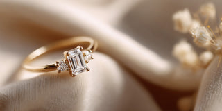 A Réia yellow gold three-stone ring with a central Emerald-cut lab-grown diamond and two round lab-grown diamond side stones, resting on beige fabric with dried flowers.