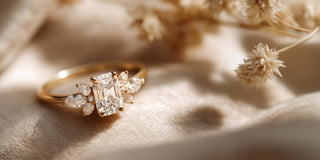 A Réia yellow gold engagement ring with an emerald cut lab-grown diamond and floral diamond side accents, resting on beige fabric with dried flowers.