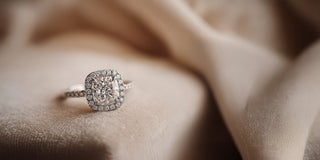 A Réia white gold engagement ring featuring a Cushion-cut lab-grown diamond with a diamond halo and pavé band, resting on beige fabric.