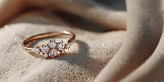 A Réia rose gold ring with a cluster of round lab-grown diamonds, resting on beige sand and fabric.