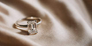 A Réia white gold solitaire ring with a Emerald-cut lab-grown diamond, resting on beige fabric.