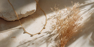 A thin gold chain necklace with five bezel-set lab-grown diamonds rests on a stone slab next to dry grass.